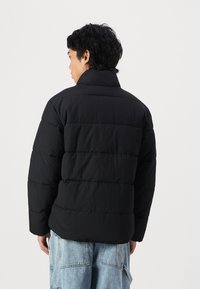 Black padded jacket with a high collar, featuring horizontal quilted design and a relaxed fit. Worn over light blue denim jeans.