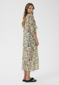Robe longue et transparente en crème avec un motif de feuilles abstraites noir et marron. Elle dispose d'un détail à nouer à la taille et de manches amples.
