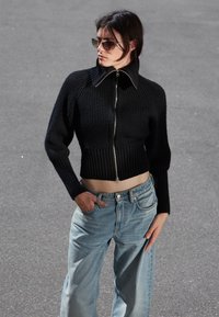 Schwarzer gerippter cropped Pullover mit hochgeschlagenem Kragen und silbernem Reißverschluss; kombiniert mit hellblauen weit geschnittenen Jeans.