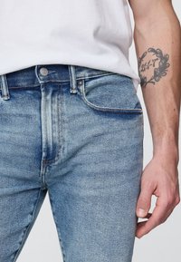 Világoskék farmernadrág karcsúsított szabással, elöl zsebekkel, fém gombbal és kontrasztos varrással. Látható textúra és kopásminták.