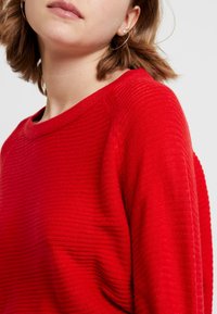 Roter Strickpullover mit gerippter Textur, weitem Ausschnitt und leicht abgesenkten Schultern. Der Stoff wirkt weich und leicht.