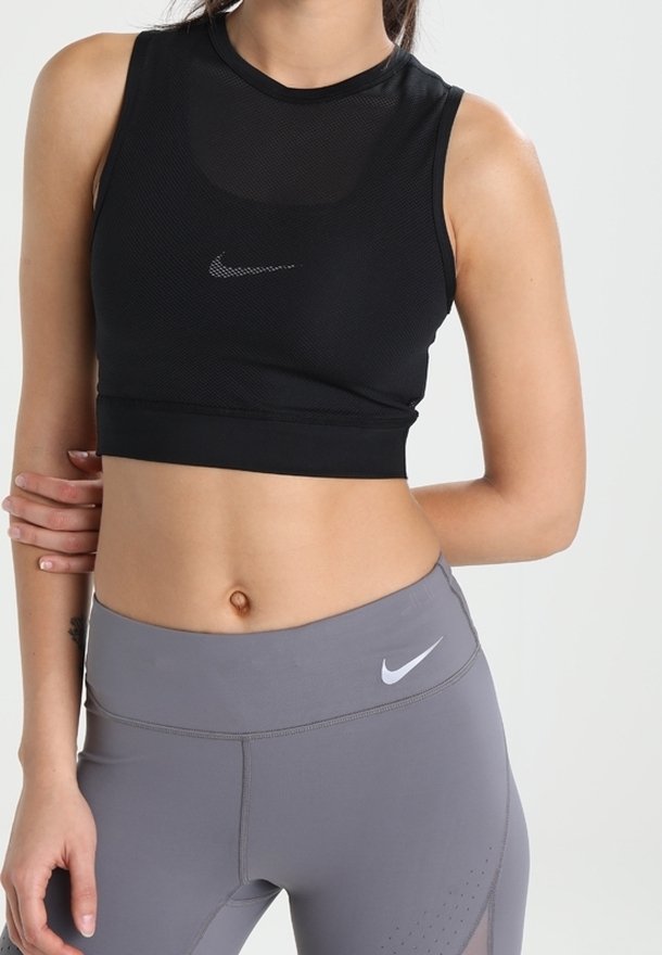 Femme portant un crop top noir sans manches et un legging de sport gris taille haute avec des logos Nike, posant avec un bras croisé sur la taille.