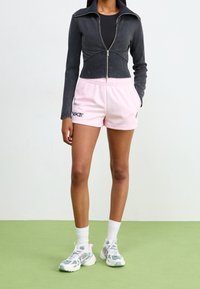 Černá zkrácená zipová bunda, růžové šortky se stahovací šňůrkou a logem, bílé ponožky, šedé a růžové tenisky. Pozadí na zelené podlaze.