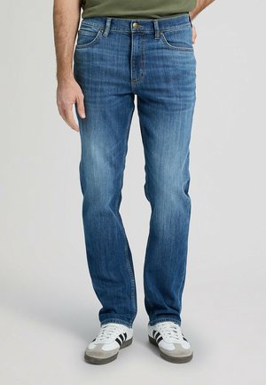 Mężczyzna ubrany w niebieskie dżinsy, oliwkowo zieloną koszulę i białe trampki z czarnymi paskami, stojący z lewą ręką swobodnie opuszczoną wzdłuż ciała.