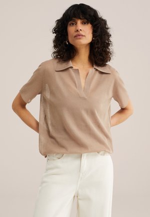 Lokkis tume juustega naine, kes kannab beeži lühikeste varrukatega polo särki ja valgeid pükse, seisab käed selja taga tühja tausta ees.