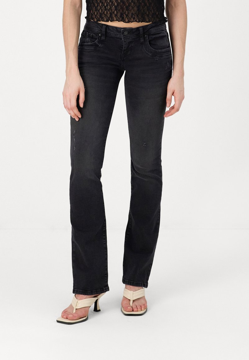 Schwarze Denim-Jeans im Schlagstil mit zerstörten Details, vorderen Taschen und mittelhoher Taille. Model trägt beigefarbene Flip-Flops mit Absätzen.