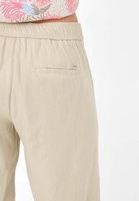 Pantalons légers en tissu stretch beige avec une taille élastique. Comprend une poche arrière unique avec un détail métal.