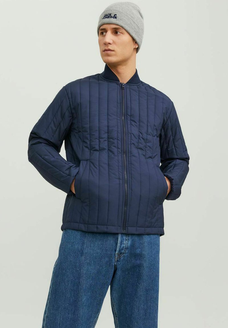 Giacca trapuntata blu navy con zip frontale, colletto alto e tasche laterali. Indossata sopra jeans a gamba larga di un blu chiaro e un berretto a maglia grigio.
