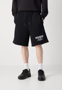 Black cotton shorts with an elastic waistband and drawstring, featuring white "HUGO" logo print on the left leg. Paired with black shoes and white socks.