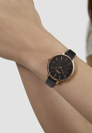 Black analog wristwatch with gold accents, round case, and a leather band. The face features minimalistic hour markers and a smooth texture.