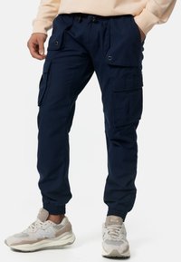 Marine cargobroek met een relaxte pasvorm, meerdere zakken, elastische boorden en een trekkoord in de taille, gecombineerd met lichtgekleurde sneakers.