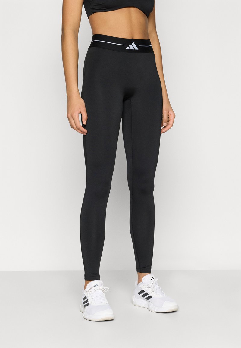 Collants noirs en matière extensible avec une taille haute ornée d'une bande blanche et d'un logo. Associés à des chaussures de sport blanches.