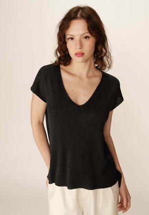 Top en maille côtelée noire avec un décolleté en V, des manches courtes, une coupe ample et des fentes sur les côtés, associé à un pantalon de couleur claire.