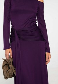 Mujer con un vestido púrpura ajustado de manga larga y detalle fruncido en la cintura, sosteniendo un bolso de mano de textura marrón con estampado.