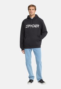 Felpa nera con un grande logo bianco "SPYDER", tasca a marsupio frontale, cappuccio con coulisse, abbinata a jeans azzurri e sneakers nere.