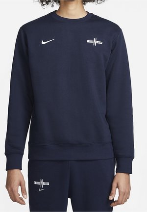 Osoba ubrana w granatową bluzę z długim rękawem oraz dopasowane spodnie z logo Nike i białym napisem "ENG" na klatce piersiowej i udzie.