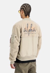Chaqueta bomber beige con un cuerpo de forro polar texturizado y mangas suaves. Presenta un logo en la espalda, puños de canalé y un bolsillo con cremallera.