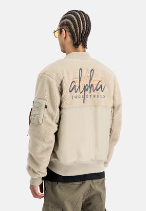 Beige Bomberjacke mit einem strukturierten Fleece-Körper und glatten Ärmeln. Verfügt über ein Logo auf der Rückseite, gerippte Bündchen und eine Reißverschlusstasche.