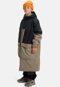 Jongere in een lange zwart en beige gewatteerde jas met capuchon, oranje muts, zwarte broek en bruine sneakers, staande tegen een witte achtergrond.