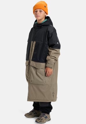 Jeune personne portant un long manteau à capuche noir et beige, un bonnet orange, un pantalon noir et des baskets marron, se tenant contre un fond blanc.