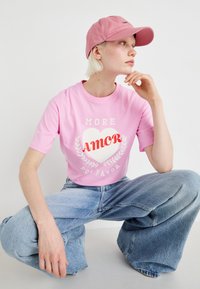 Růžové tričko s krátkým rukávem má grafiku "MORE AMOR" srdce, které je kombinováno s volnými světle modrými džíny. Růžová čepice je nošena vpředu.
