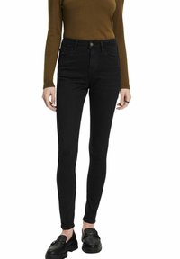 Personne portant un jean noir slim, un haut marron à manches longues et des mocassins noirs, debout sur un fond blanc.