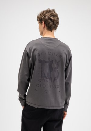Jonge man met krullend haar die een donkergrijs shirt met lange mouwen draagt met grote vervaagde zwarte grafische tekst op de achterkant, staand tegen een eenvoudige lichte achtergrond.