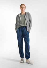 Šedá mikina na zip přes zelené tričko, doplněná volnými modrými tepláky a světlými teniskami. Hladká látka s elastickými manžetami a páskem.
