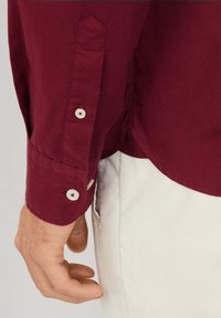 Camicia bordeaux con bottoni ai polsini, colletto appuntito e orlo curvo. Realizzata in un tessuto morbido, esposta sul braccio di una persona.