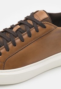 Zapatilla de cuero marrón con cordones marrones oscuros y una suela de goma texturizada. Presenta costuras en contraste y un logo de Geox en el lateral.