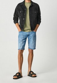 Chaqueta de mezclilla negra, camiseta verde oliva, pantalones cortos azul claro y sandalias negras con suelas marrones; conjunto casual con colores contrastantes.