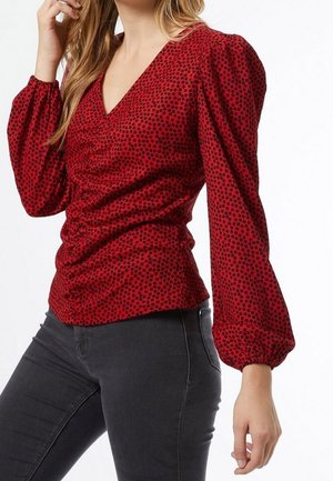 Žena oblečená v červené haleně s dlouhými rukávy a vzorem, s nařaseným předním dílem, a černých přiléhavých džínách, pózující s jednou rukou u obličeje.