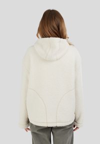 Kvinde med langt hår iført en cremefarvet fleece hættetrøje og grå bukser, vist fra ryggen på en ensfarvet baggrund.