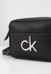 Sort syntetisk læder skuldertaske med en glat overflade, der har et fremtrædende sølv "ck" logo og en justerbar stropp.