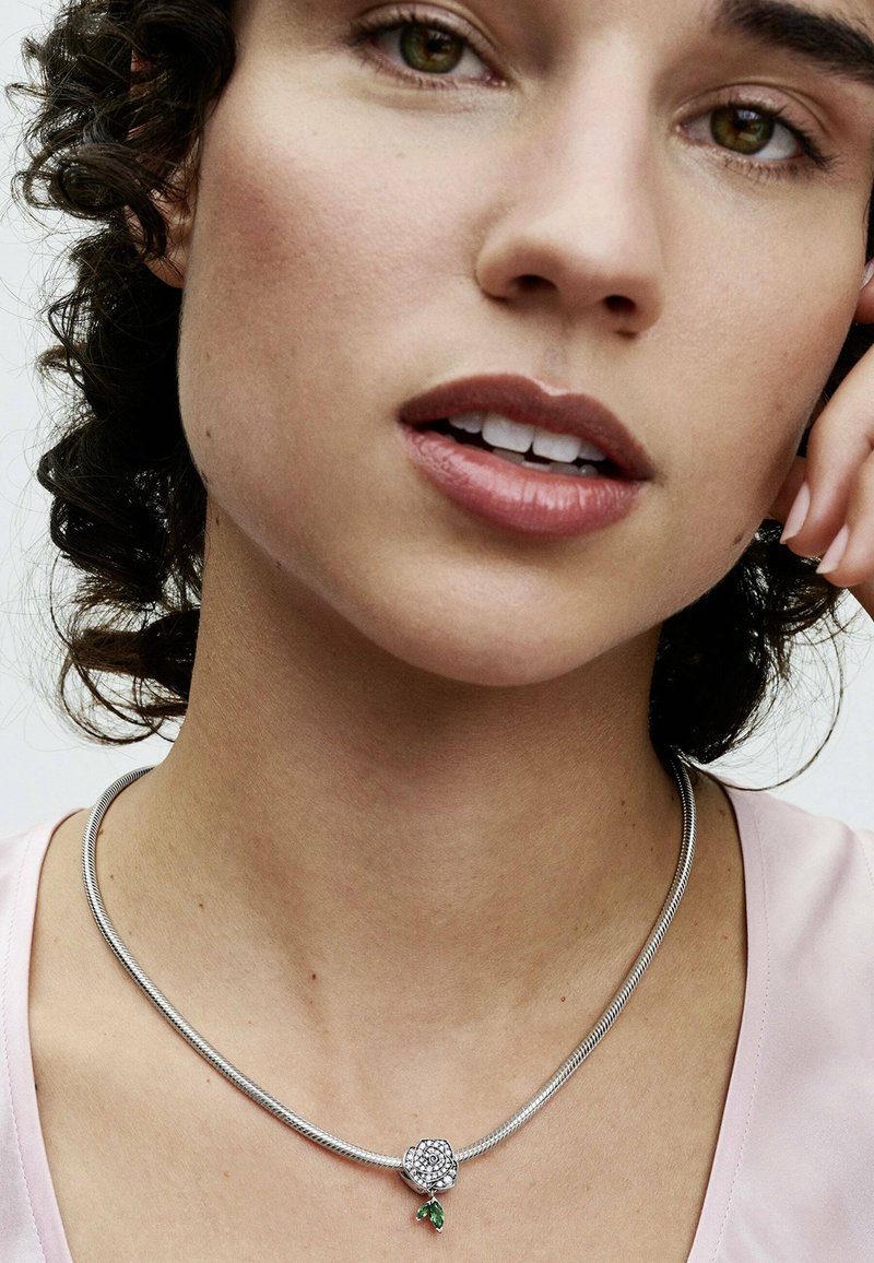 Silberne Schlangenkette mit einem Blumenanhänger, der funkelnde Steinakzente und ein grünes Blätterndetail aufweist. Texturiertes und modernes Design.