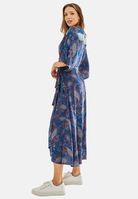 Blue paisley patterned dress with a tied waist and three-quarter sleeves; paired with white sneakers. Smooth fabric and ankle-length design.