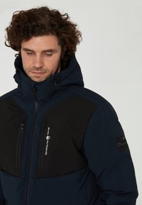 Marinblå isolerad jacka med svarta paneler, dragkedjefickor och huva. Har en logotyp på bröstet och strukturerade tygdetaljer.