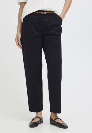 Vrouw draagt zwarte, op maat gemaakte enkellange broek met zijzakken, een bruine riem, witte top en zwarte Mary Jane-schoenen.