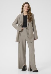 Woman with long wavy hair wearing a beige suit with wide-leg pants and a black shirt, smiling and looking to the side against a white background.