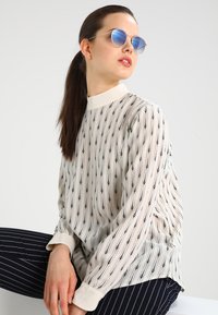 Blusa a maniche lunghe in tessuto crema con strisce verticali nere e motivi a rombi. Presenta un colletto alto e polsini con bottoni.