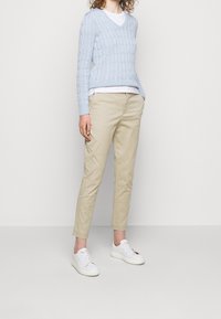 Svetle modrý svetr s pleteným vzorem, s výstřihem do V; kombinovaný s béžovými chino kalhotami. Bílé tenisky dodávají kontrast. Jednoduchý a ležérní outfit s čistými liniemi.