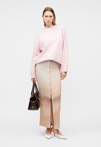 Femme portant un pull rose clair et une jupe longue beige boutonnée, tenant un sac à main marron foncé et chaussant des talons blancs.