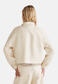 Krátká béžová bunda s límcem, bočními kapsami a dlouhými rukávy. Hladká látka s minimálními švy vzadu.