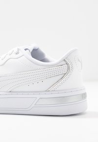 Bílé sportovní boty s hladkým syntetickým svrškem, stříbrnými akcenty, texturovanou podešví a šněrovacím designem. Obsahují logo na patě a perforované detaily.