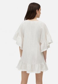 Vestido de lino blanco con mangas y dobladillo ondulados. Corte holgado y textura lisa, presenta un diseño sencillo en la parte posterior y un corte a la altura de la rodilla.