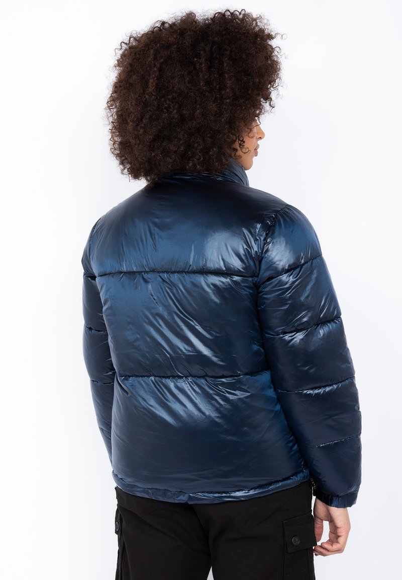 Veste matelassée bleu marine avec un fini brillant et matelassé, dotée d'un col haut et de motifs en coutures horizontales. Design léger et isolant.
