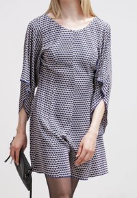 Námořnické a bílé vzorované šaty s křidélkovými rukávy, kulatým výstřihem a krátkým střihem. Modelka drží černou kabelku.