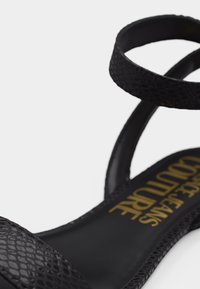 Μαύρο επίπεδο σανδάλι με υφασμένες λουρίδες και χρυσό ανάγλυφο όνομα επωνυμίας στη σόλα, απεικονίζεται σε λευκό φόντο.