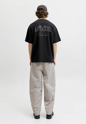 Person wearing a black cap, black T-shirt with "The True Apparel" text, light gray loose pants, and black shoes, standing facing away on white background.
