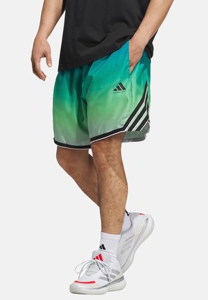  Uomo che indossa pantaloncini da basket sfumati verdi e sneaker Adidas bianche, in piedi con una mano in tasca.